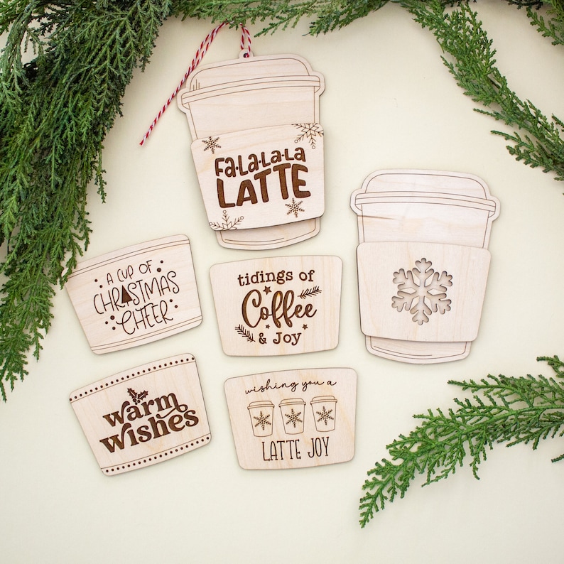 May include: Wooden Christmas ornaments in the shape of coffee cups and cup sleeves. The ornaments have engraved text such as "Fa-La-La-La LATTE", "A cup of Christmas Cheer", and "Warm Wishes". The ornaments are decorated with snowflakes and other festive designs.
