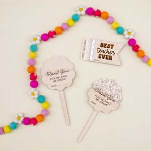 May include: Wooden teacher appreciation gifts, including a sign that reads "Best Teacher Ever" and two flower-shaped signs that say "Thank You For Helping Me Grow." A colorful felt ball garland with daisy accents is also present.