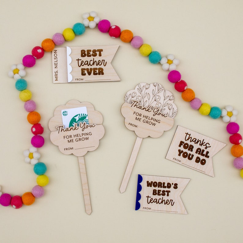May include: A collection of wooden teacher appreciation gifts. The gifts include flower-shaped gift card holders, and banner-shaped tags with phrases like "Best Teacher Ever" and "Thanks for all you do." A colorful felt ball garland is in the background.