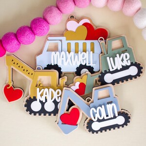 May include: Wooden construction vehicle name tags in yellow, blue, and green, each with a child's name in white. The tags include a dump truck, excavator, and bulldozer, with heart accents. A pink and white felt ball garland is in the background.