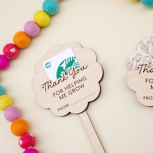 May include: Wooden flower-shaped gift card holders with the text "Thank You For Helping Me Grow" and a space for a name. A Starbucks gift card is in one holder. A colorful felt ball garland is in the background.