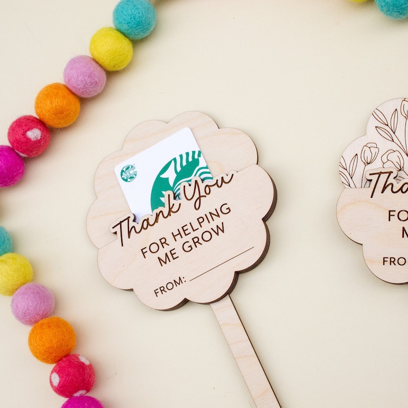 May include: Wooden flower-shaped gift card holders with the text "Thank You For Helping Me Grow" and a space for a name. A Starbucks gift card is in one holder. A colorful felt ball garland is in the background.