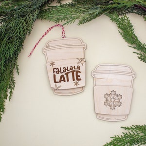 May include: Two wooden coffee cup ornaments. One ornament has the text "Fa-La-La-La LATTE" and snowflake designs. The other ornament has a snowflake design. Both ornaments have a light wood finish and are hanging with red and white twine.