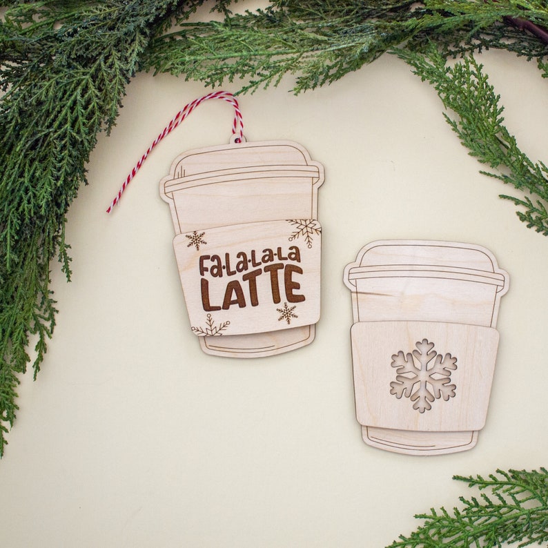 May include: Two wooden coffee cup ornaments. One ornament has the text "Fa-La-La-La LATTE" and snowflake designs. The other ornament has a snowflake design. Both ornaments have a light wood finish and are hanging with red and white twine.