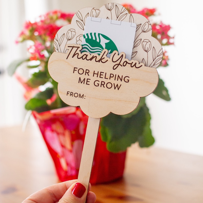 May include: A wooden plant stake with a cloud-shaped design and the words "Thank You For Helping Me Grow" printed on it. The stake holds a gift card with a green logo. The background features a red flower pot and pink flowers.
