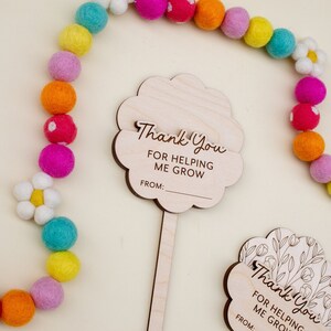 May include: Wooden flower-shaped thank you cards with the text "Thank You FOR HELPING ME GROW" and a space for a name. A colorful felt ball garland and a white daisy are in the background.