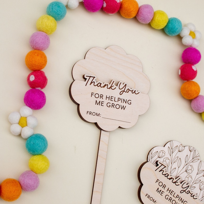 May include: Wooden flower-shaped thank you cards with the text "Thank You FOR HELPING ME GROW" and a space for a name. A colorful felt ball garland and a white daisy are in the background.