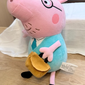May include: A plush toy of a pink pig character wearing glasses, a light blue shirt with a white collar and blue tie, and black shoes. The character holds a small mustard-yellow satchel in one hand and has a white fabric tag attached to its side.
