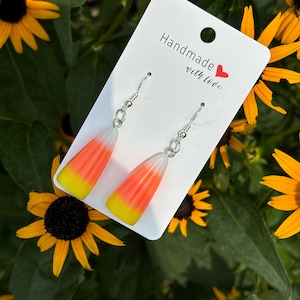 Pode incluir: Um par de brincos pendentes com um design de milho doce. Os brincos são feitos de argila polimérica branca, laranja e amarela e são presos a ganchos prateados. Os brincos são exibidos em um cartão branco com o texto "Handmade with love".
