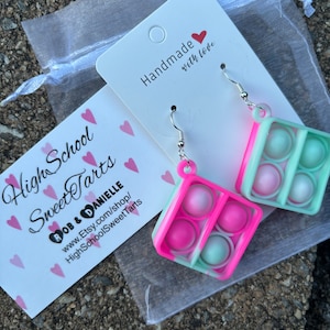 May include: A pair of pink and green pop it earrings with silver hooks. The earrings are handmade with love and are packaged in a white organza bag with a pink and white business card that says "High School Sweet Tarts" and "Bob & Danielle".