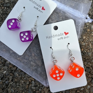 May include: Two pairs of dangle earrings with dice charms. One pair has purple dice charms and the other pair has orange dice charms. The earrings are on white cards with the text "Handmade with love" on each card.