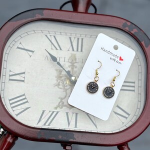 May include: A pair of gold-colored earrings featuring a black clock face design. The earrings are displayed on a white card with the text "Handmade with love" and a red heart. The background includes a vintage-style clock.