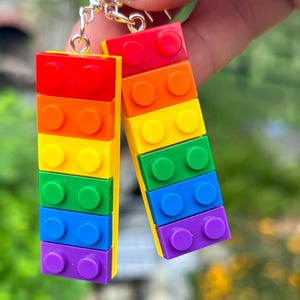 Puede incluir: Un par de pendientes coloridos hechos de bloques rectangulares apilados en un esquema de color arcoíris. Los pendientes presentan bloques rojos, naranjas, amarillos, verdes, azules y morados, conectados con ganchos plateados.
