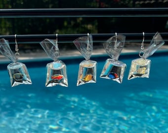 May include: Five pairs of dangle earrings with small plastic bags containing colorful fish. The earrings are hanging from a black metal bar against a blue pool background.