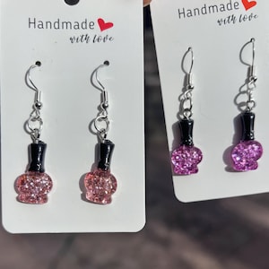Könnte beinhalten: Zwei Paar Hängeohrringe mit einem pinkfarbenen Glitzer-Nagellackflaschen-Anhänger. Die Ohrringe sind silberfarben und haben einen Hakenverschluss. Die Karte hinter den Ohrringen lautet "Handmade with love".