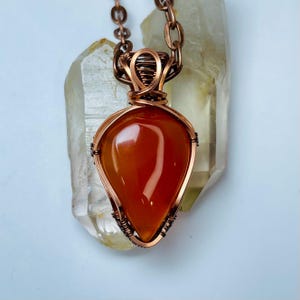 Könnte beinhalten: Handgefertigte Kupfer-Anhänger-Halskette mit einem tropfenförmigen, orange-roten Edelstein. Der Stein ist in eine Kupferdrahtwicklung eingefasst und an einer passenden Kupferkette aufgehängt. Der Anhänger ist ca. 3,8 cm lang.