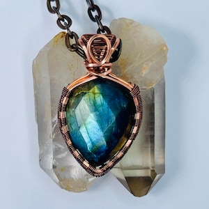 May include: A copper wire wrapped pendant featuring a blue-green labradorite gemstone. The pendant is displayed on top of two clear quartz crystals.