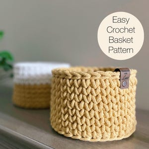 May include: Two handmade crochet baskets. The front basket is mustard yellow with a leather tag that says "Handmade with Love". The second basket is white and mustard yellow. The image also includes the text "Easy Crochet Basket Pattern".