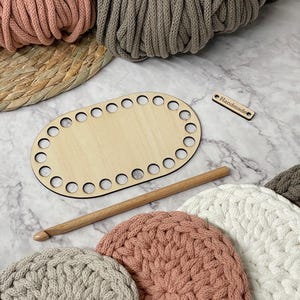 May include: A wooden oval basket base with pre-drilled holes, a wooden crochet hook, and several crocheted circles in shades of pink, gray, and white. The word "Handmade" is on a small wooden tag. Rolls of yarn are in the background.