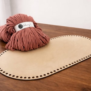 May include: A large oval-shaped light-colored wooden base featuring a series of evenly spaced holes along its perimeter, paired with a thick skein of dusty rose-colored cord wrapped in a white paper label with the brand name Bobbiny printed on it.