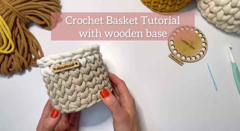 Op de afbeelding: Een handgemaakte gehaakte mand met een houten bodem. De mand is beige met een witte rand en een klein houten label met de tekst "handmade". De afbeelding toont ook haakbenodigdheden en de tekst "Crochet Basket Tutorial with wooden base".