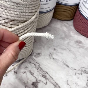 May include: Close-up of a hand holding a piece of off-white macrame cord, with a frayed end. Several spools of macrame cord in various colors are in the background, with labels that read "crame SUPPLIES". The cord is likely used for crafting projects.