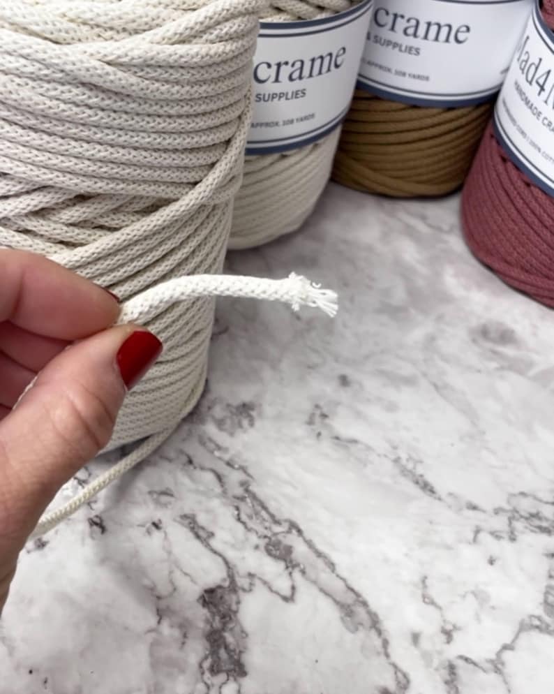 May include: Close-up of a hand holding a piece of off-white macrame cord, with a frayed end. Several spools of macrame cord in various colors are in the background, with labels that read "crame SUPPLIES". The cord is likely used for crafting projects.
