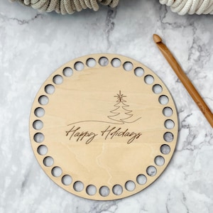 May include: A round, light-colored wooden base with evenly spaced holes around the edge. The base features a laser-etched Christmas tree and the words "Happy Holidays" in a script font. A wooden crochet hook rests on the side.
