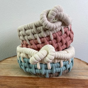 May include: Three woven baskets in beige, pink, and blue, stacked on top of each other. The baskets are made of a thick rope and have a knotted handle.