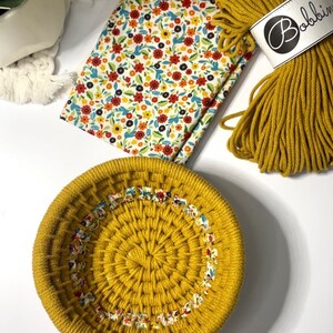 May include: A round, mustard yellow macrame basket with a floral fabric trim. The basket is made with thick cotton cord and is sitting on a white surface.