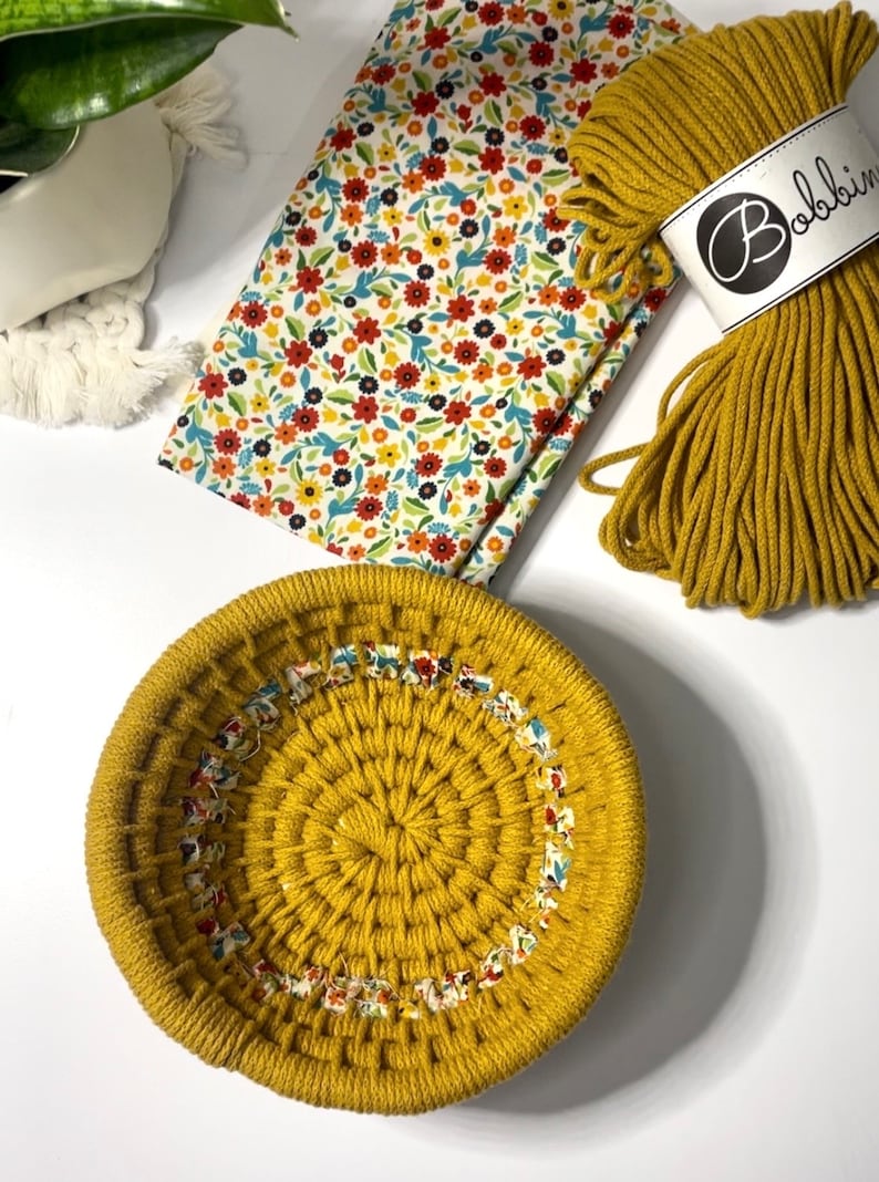 May include: A round, mustard yellow macrame basket with a floral fabric trim. The basket is made with thick cotton cord and is sitting on a white surface.