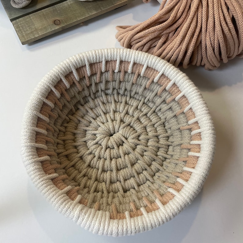 May include: A round, woven basket with a white and peach coloured border. The basket is made of natural fibres and has a circular, textured pattern.