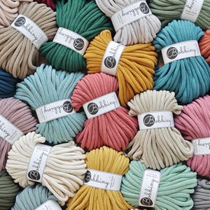 May include: A collection of colorful Bobbiny jumbo cotton yarn bundles. The yarn is available in various colors, including teal, pink, yellow, and beige. Each bundle is wrapped with a white label featuring the Bobbiny logo.