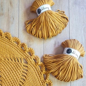 Puede incluir: Dos carretes de cuerda de algod&oacute;n amarilla mostaza, cada uno etiquetado con "Bobbiny", con una alfombra de crochet amarilla mostaza en el fondo.