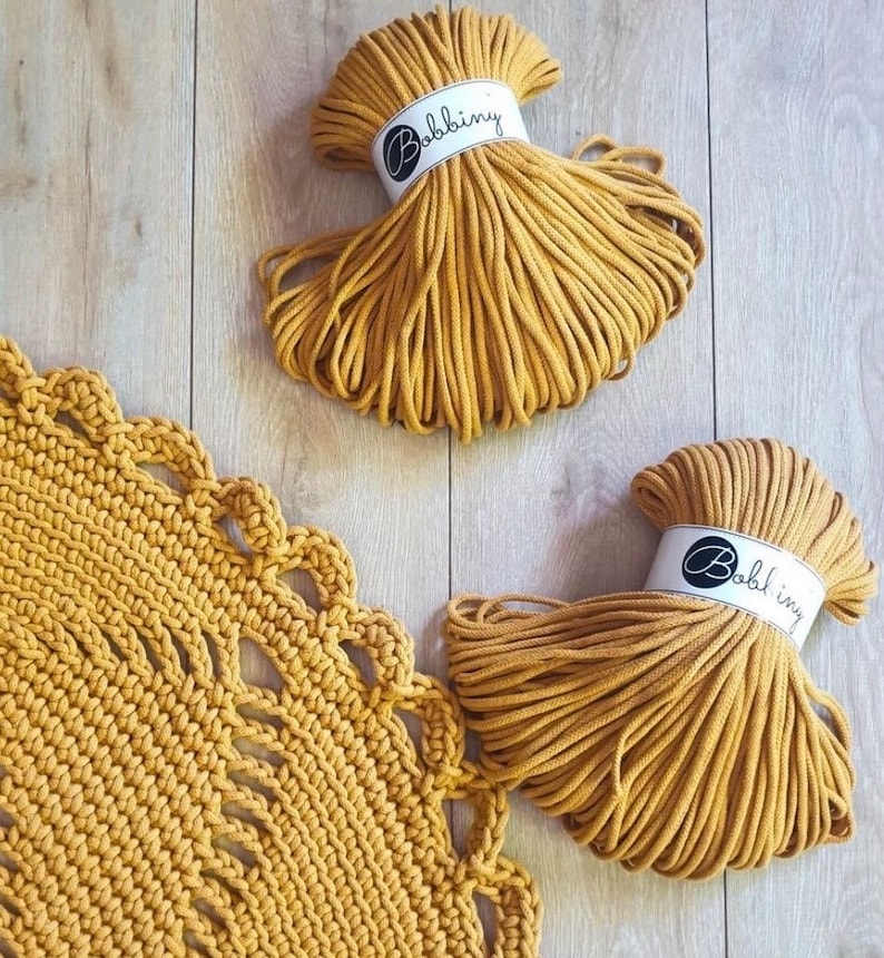 Puede incluir: Dos carretes de cuerda de algod&oacute;n amarilla mostaza, cada uno etiquetado con "Bobbiny", con una alfombra de crochet amarilla mostaza en el fondo.