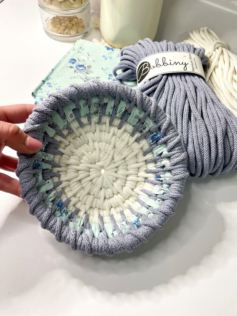 May include: A close-up of a grey and white crocheted coaster with a blue and white floral pattern. The coaster is made with a thick, soft yarn and has a textured surface. The text "Bobbiny Premium Cotton String" is visible on the yarn.