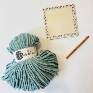 May include: A craft kit featuring a ball of teal-colored yarn with a "Bobbiny" label, a wooden crochet hook, and a square wooden base with evenly spaced holes around the perimeter. The items are arranged on a white surface.