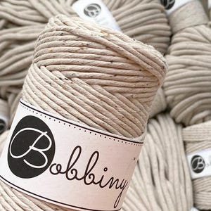 May include: Close-up of a beige cotton rope with a white label that says "Bobbiny". The rope is wound into a ball shape.