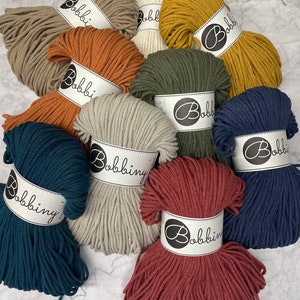 Puede incluir: Surtido de 10 ovillos de cuerda de algod&oacute;n en varios colores, incluyendo beige, marr&oacute;n, verde, azul y rojo. Cada ovillo tiene una etiqueta blanca con el texto "Bobbiny" impreso en ella.
