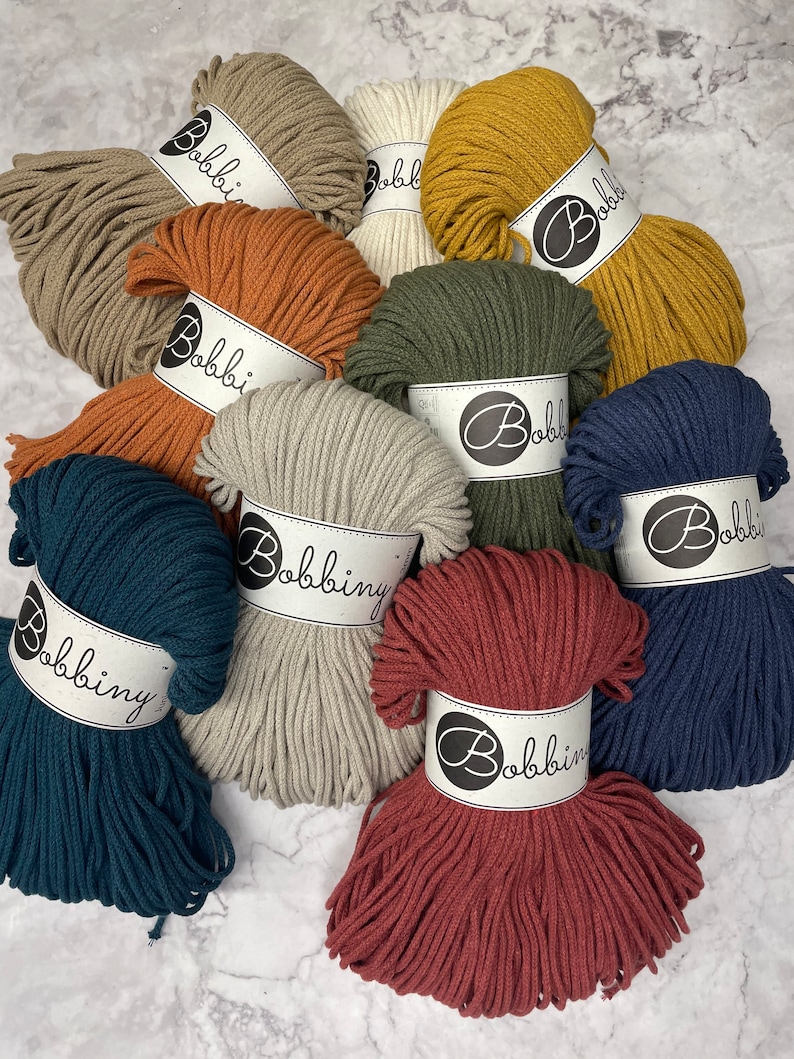Puede incluir: Surtido de 10 ovillos de cuerda de algod&oacute;n en varios colores, incluyendo beige, marr&oacute;n, verde, azul y rojo. Cada ovillo tiene una etiqueta blanca con el texto "Bobbiny" impreso en ella.