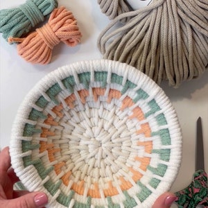 May include: A white macrame bowl with a green, peach, and white colour pattern. The bowl is being held by a person's hands. The image also shows a ball of beige rope with the label "Bobbiny" and two balls of rope in green and peach.