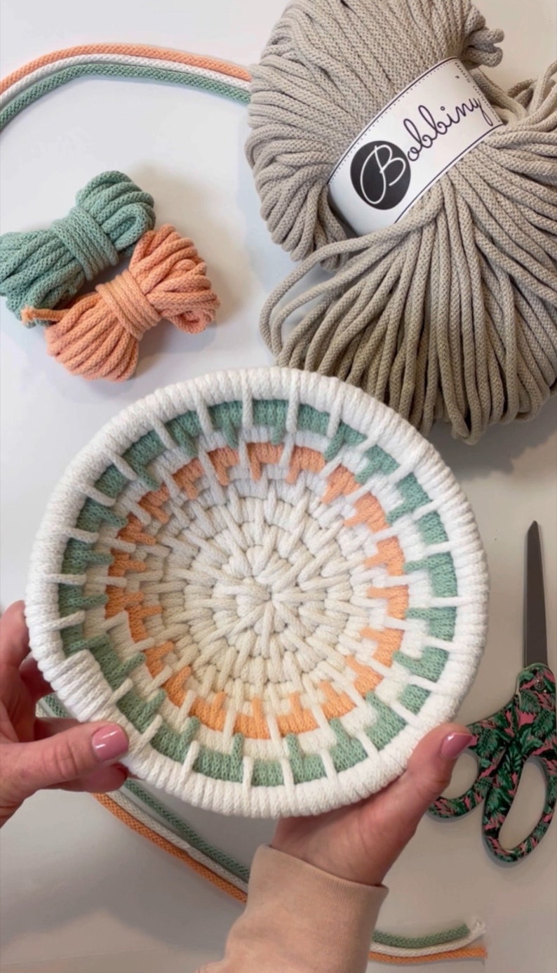May include: A white macrame bowl with a green, peach, and white colour pattern. The bowl is being held by a person's hands. The image also shows a ball of beige rope with the label "Bobbiny" and two balls of rope in green and peach.
