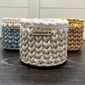 Op de afbeelding: Drie handgemaakte gehaakte manden in blauwgrijs, beige en geelbruin. Elke mand heeft een witte gevlochten rand en een klein houten label met de tekst "Handmade". De cilindrische manden staan op een donker houten oppervlak.
