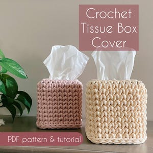 May include: Two crochet tissue box covers, one in a dusty rose color and the other in cream, sit side-by-side. The tissue boxes are square with a textured, woven appearance. White tissues peek out from the top of each box. Text reads "Crochet Tissue Box Cover" and "PDF pattern & tutorial."