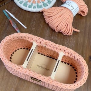 May include: A peach-colored crocheted basket with two dividers, made with cotton cord. The basket is sitting on a wooden surface. The label "Mac4Macrame" is visible on the basket.