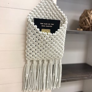 May include: A macrame wall hanging with a pocket for storing items. The hanging is made of light beige cord and has a fringe at the bottom. A book titled "The Face on the Milk Carton" is in the pocket.