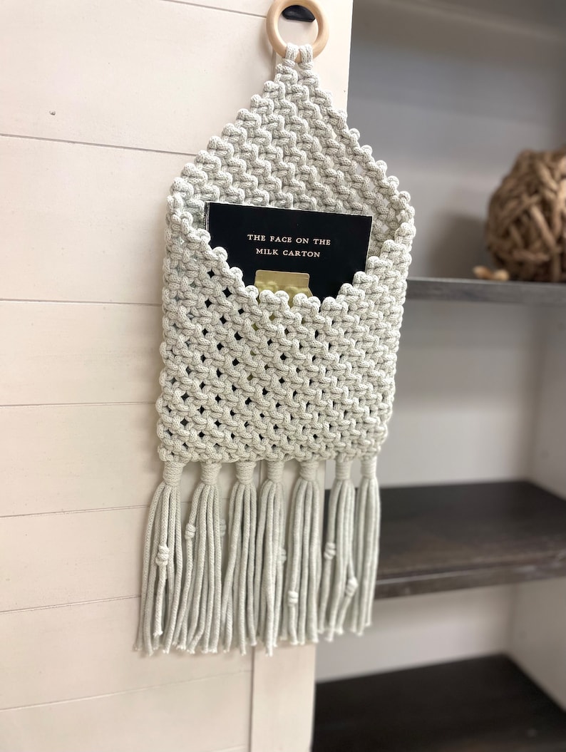 May include: A macrame wall hanging with a pocket for storing items. The hanging is made of light beige cord and has a fringe at the bottom. A book titled "The Face on the Milk Carton" is in the pocket.