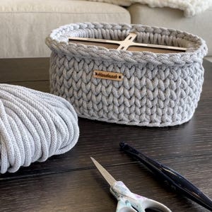 May include: A handmade gray crochet basket with a wooden base and a small "Handmade" tag. A ball of gray yarn, scissors, and a crochet hook are on the dark wooden surface. The basket is oval-shaped and appears to be sturdy.