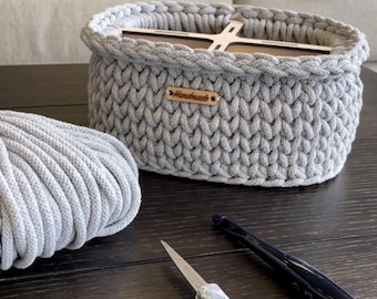 Puede incluir: Una cesta de ganchillo gris hecha a mano con base de madera y una pequeña etiqueta "Handmade". Una madeja de hilo gris, tijeras y un ganchillo están sobre la superficie de madera oscura. La cesta es de forma ovalada y parece resistente.