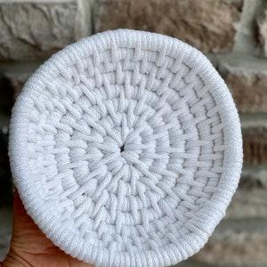 May include: A white crocheted bowl with a circular shape and a woven pattern. The bowl is made of thick white rope and has a smooth, even texture.